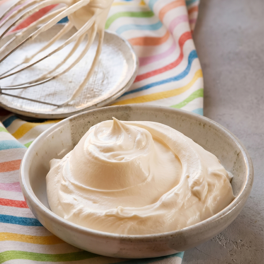 A large mixing bowl filled with fluffy white whipped cream frosting on a colorful striped cloth, with a whisk resting inside.