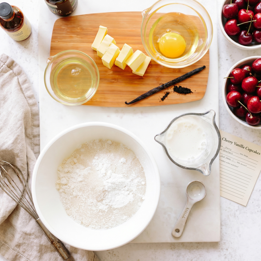 Vanilla cupcake ingredients arranged on a kitchen counter with flour, sugar, butter, eggs, milk, and cherries