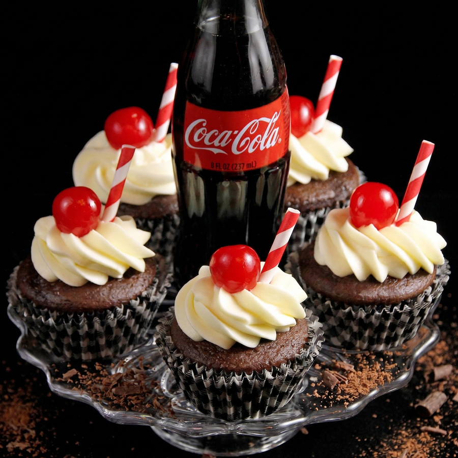 Coca-Cola Cupcakes Five chocolate cupcakes topped with white frosting, red cherries, and striped straws, arranged on a glass plate next to a bottle of Coca-Cola.