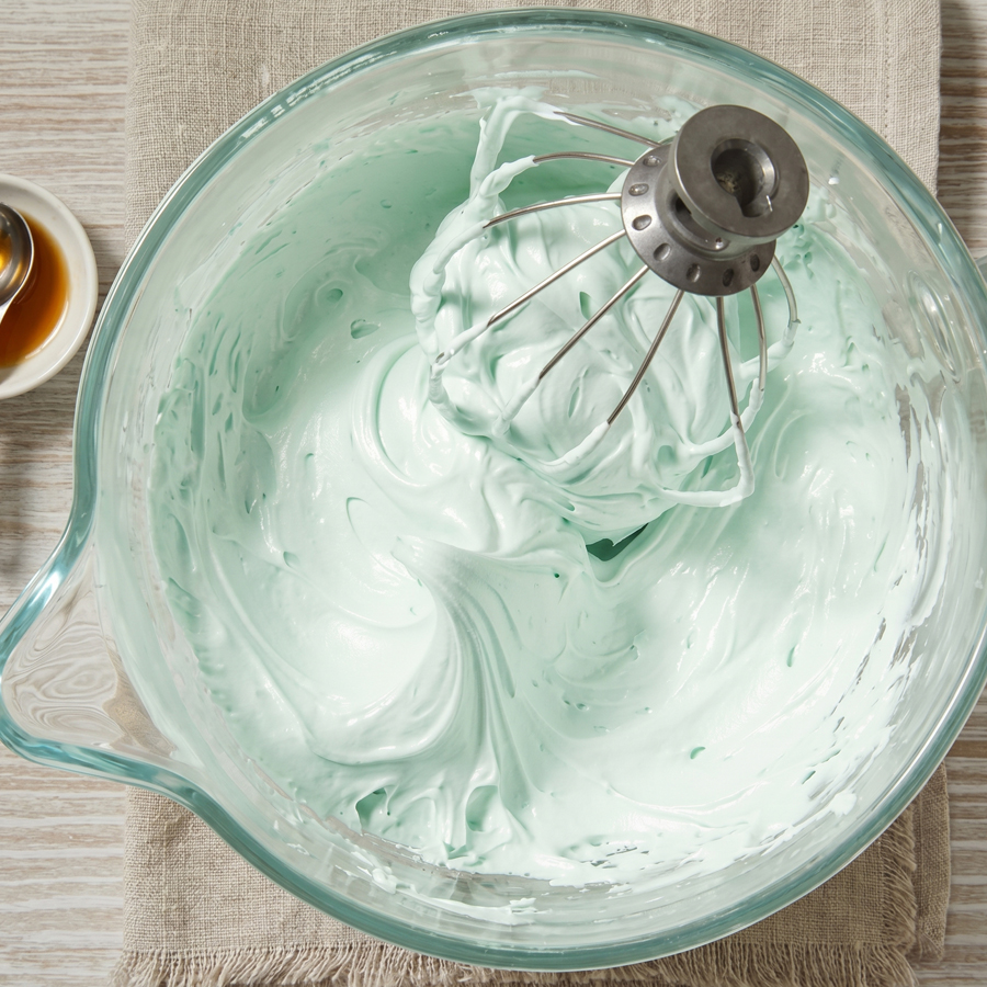 Glossy seafoam frosting in a glass bowl with a spoon lifting a thick strand