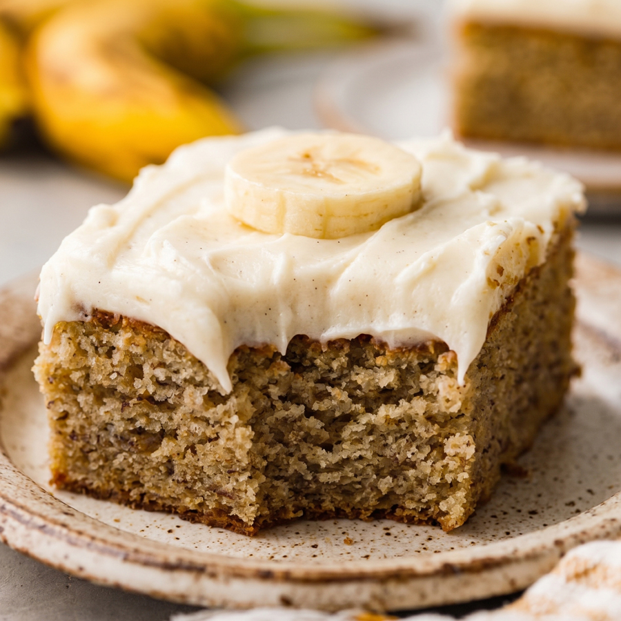 A square slice of banana cake topped with a thick layer of cream cheese frosting and fresh banana slices on a plate.