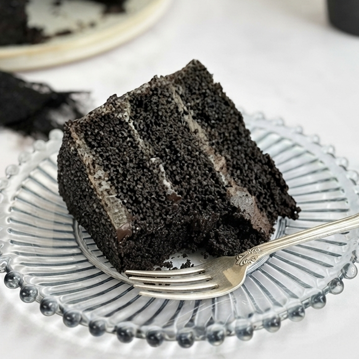 The Ultimate Black Chocolate Cake A tall slice of dark, almost black chocolate cake frosted with a smooth white vanilla buttercream on a plate.